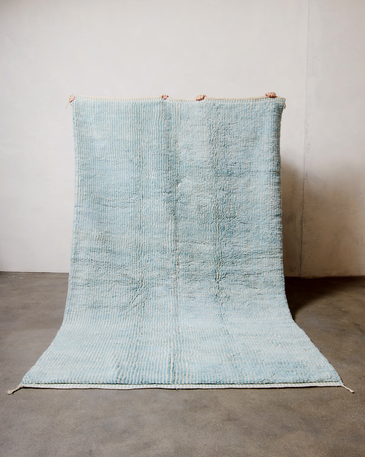 Berberlin Beni M'rirt rug in cream and light blue stripes being held up against a white wall in a studio.